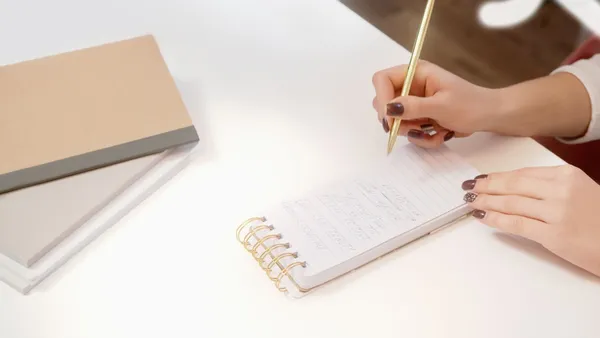 Person writing with a pencil in a spiral notebook at a desk.