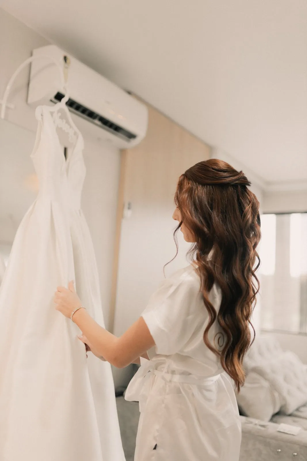 Woman in a robe looking at a wedding dress hanging in a room.