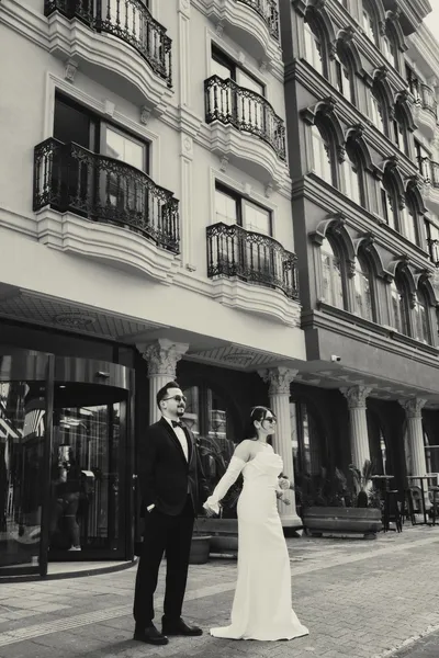Couple in wedding attire in front of a building facade, in black and white.