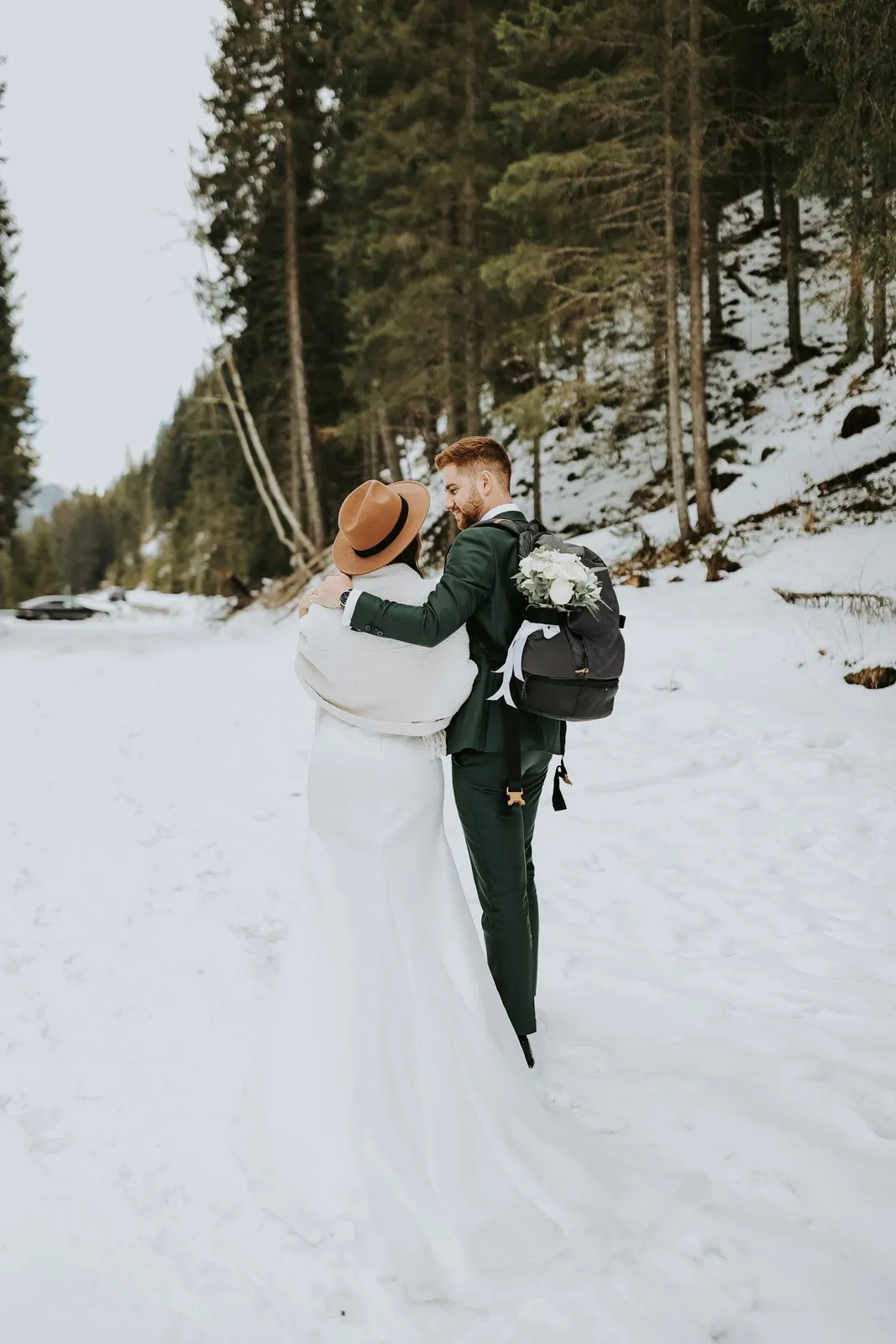 Person in a suit carrying a bride on a snowy path beside fir trees.