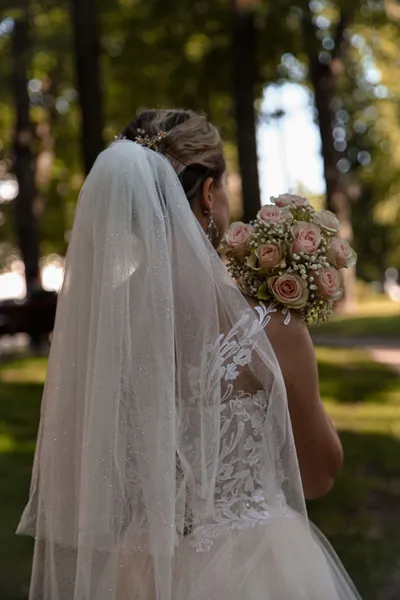 Bride seen from behind with her veil and a bouquet in a sunlit garden.