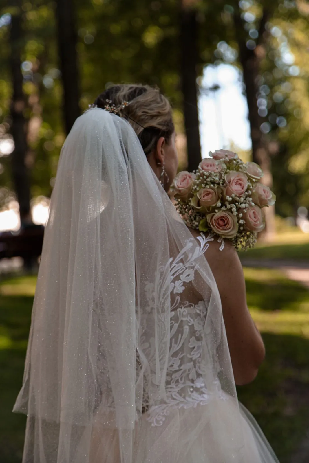 Bride seen from behind with her veil and a bouquet in a sunlit garden.