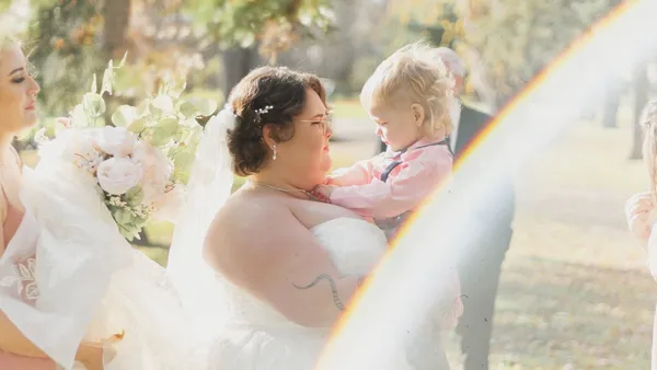 A bride in a white wedding dress holds a child during an outdoor ceremony.