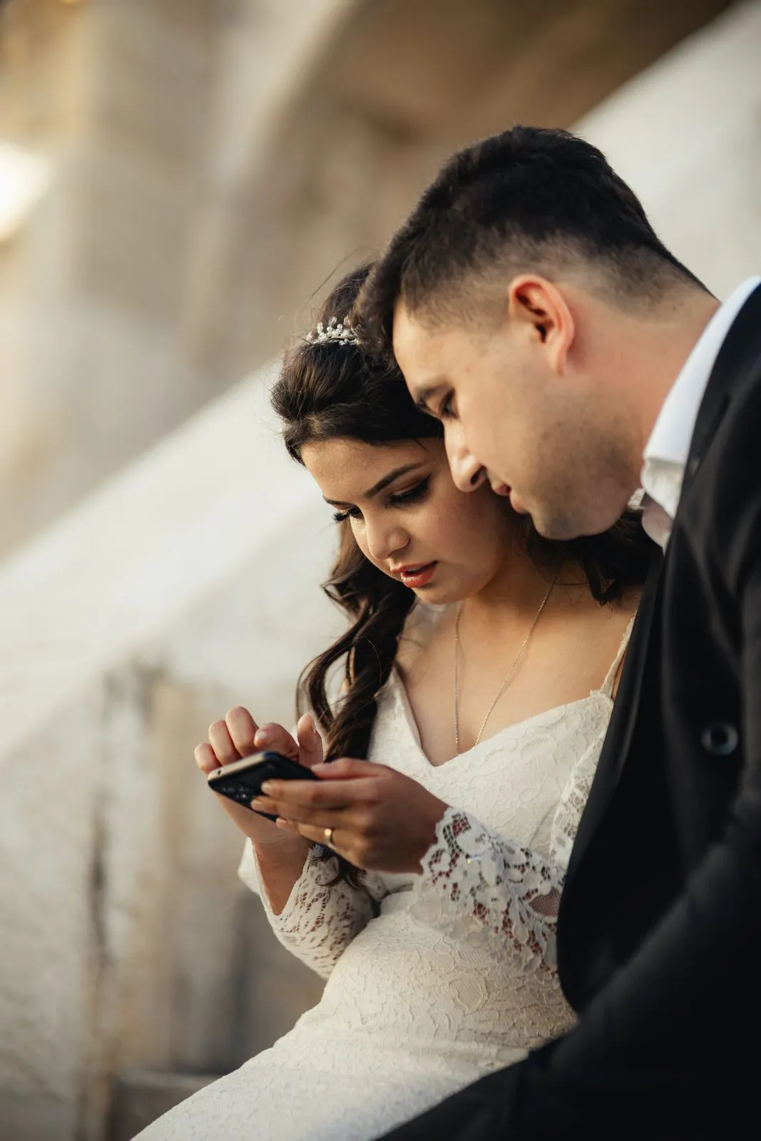 Bride and groom sitting together, using smartphone on their wedding day.
