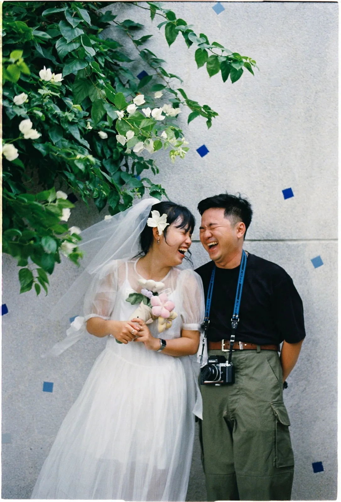 Mariée et homme portant un appareil photo autour du cou, riant ensemble devant un mur fleuri.