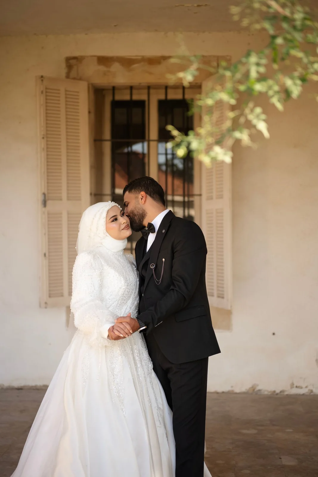 Marié embrassant la joue de la mariée, leurs mains réunies devant une fenêtre à volets.