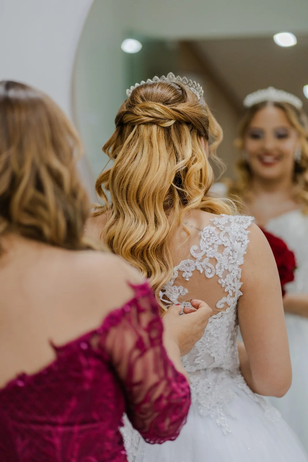 A person adjusts the back of the bride’s dress in front of a mirror.
