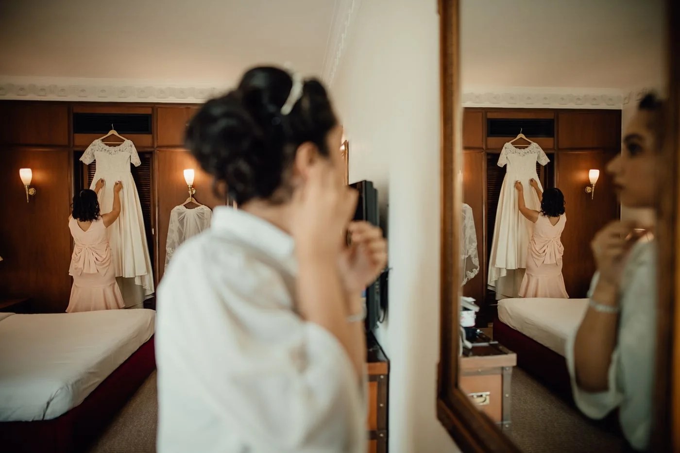 Preparations in front of a bedroom mirror with a wedding dress hanging in the background.