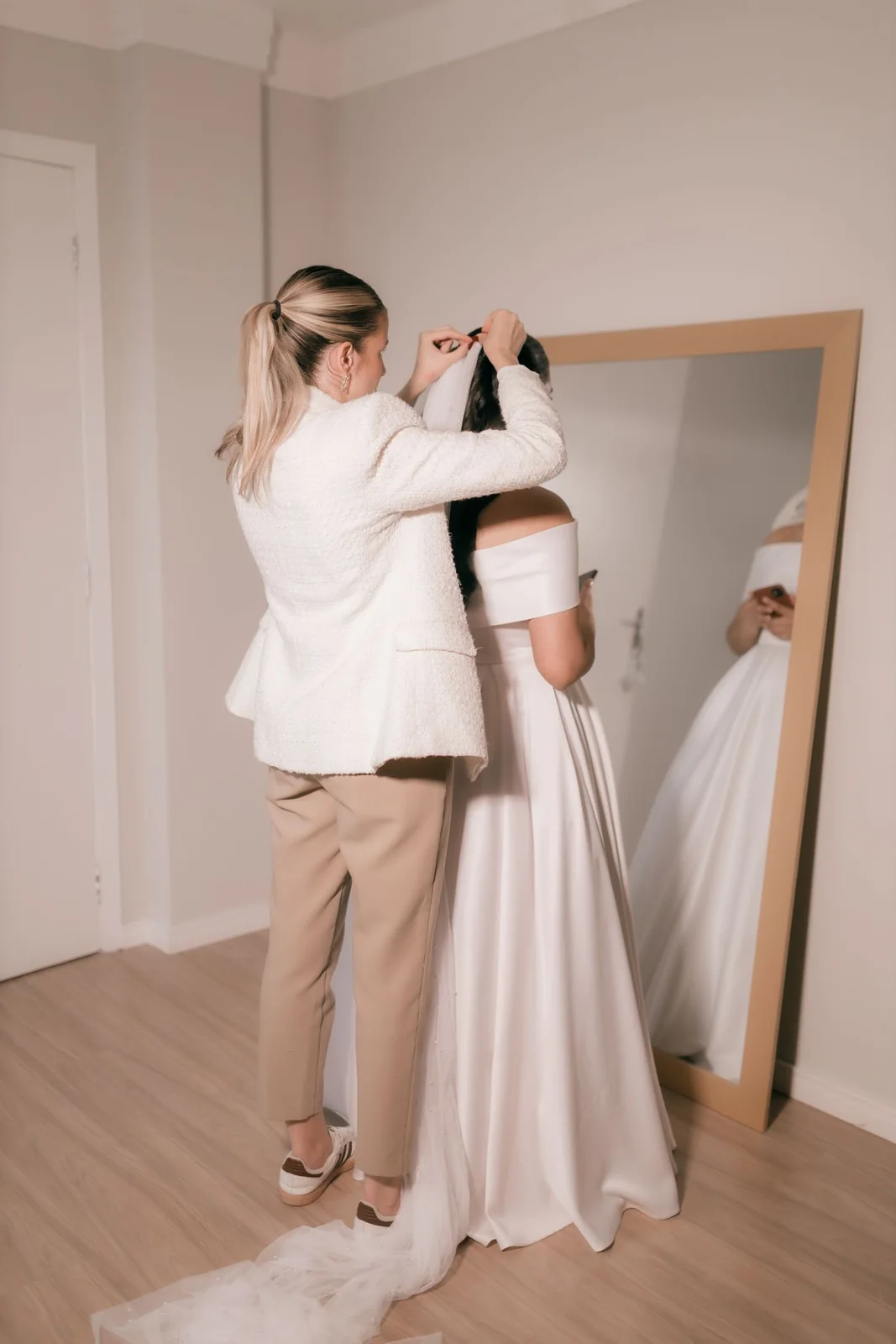 Une personne ajuste la coiffure d’une mariée en robe blanche devant un miroir.