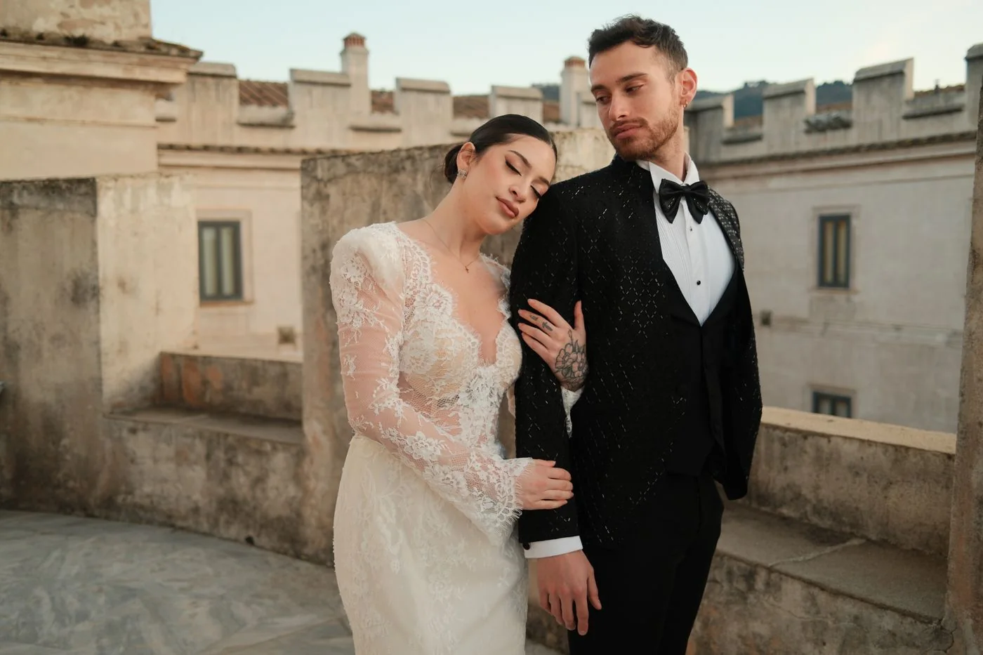 Une mariée appuie sa tête sur l’épaule du marié devant un bâtiment en pierre.
