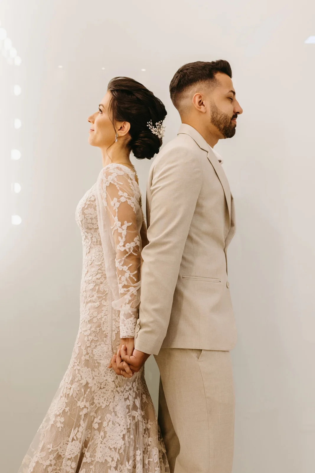 Two newlyweds hold hands while standing back to back against a pale wall.