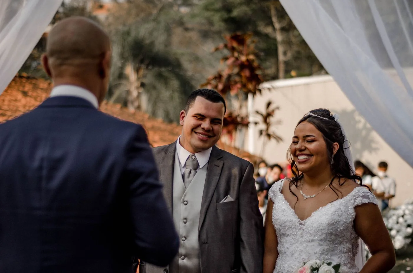 Une mariée et un marié sourient face à un officiant pendant une cérémonie extérieure.