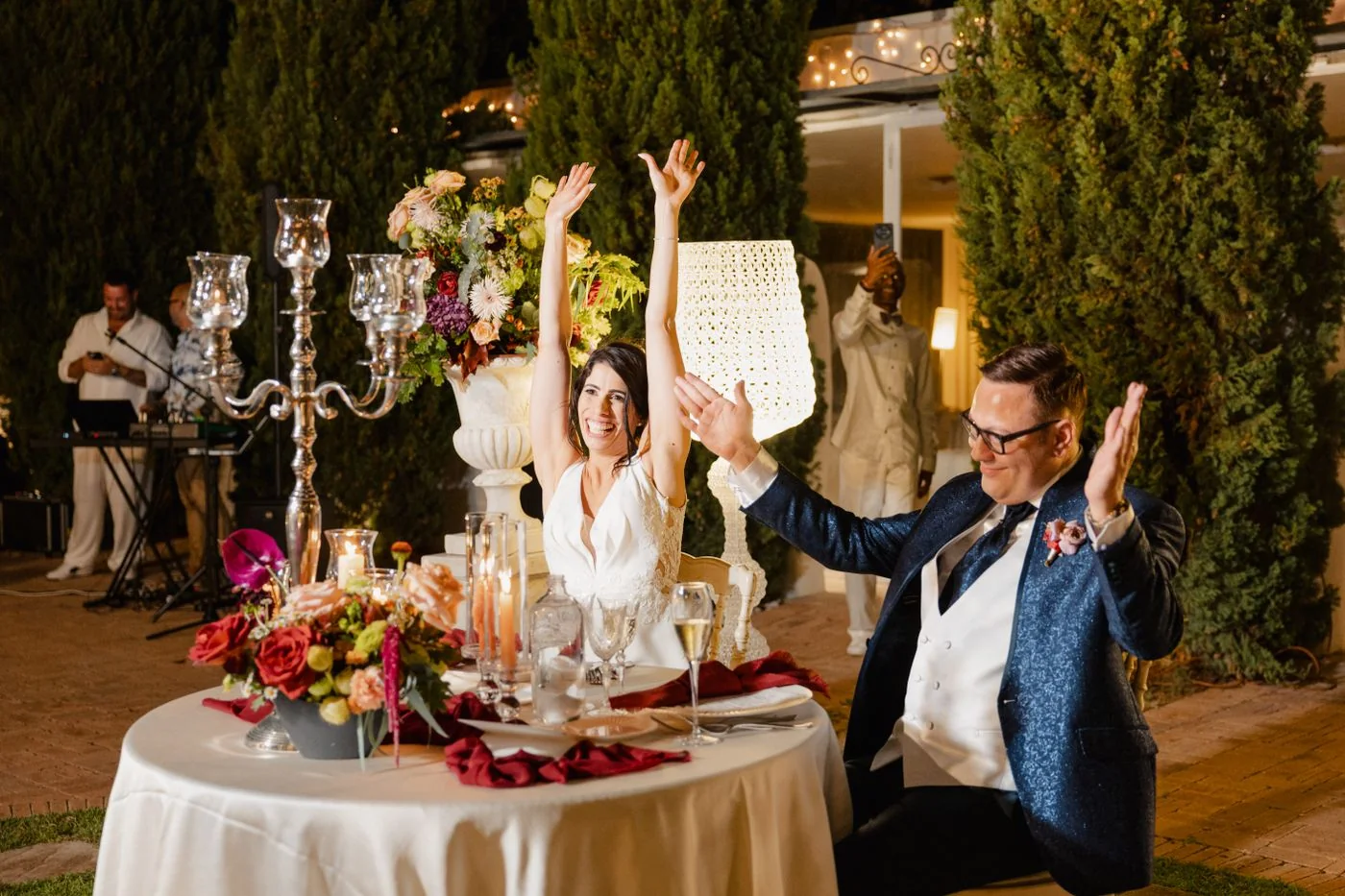 Newlyweds raise their glasses behind a flower-decorated table at a nighttime reception.