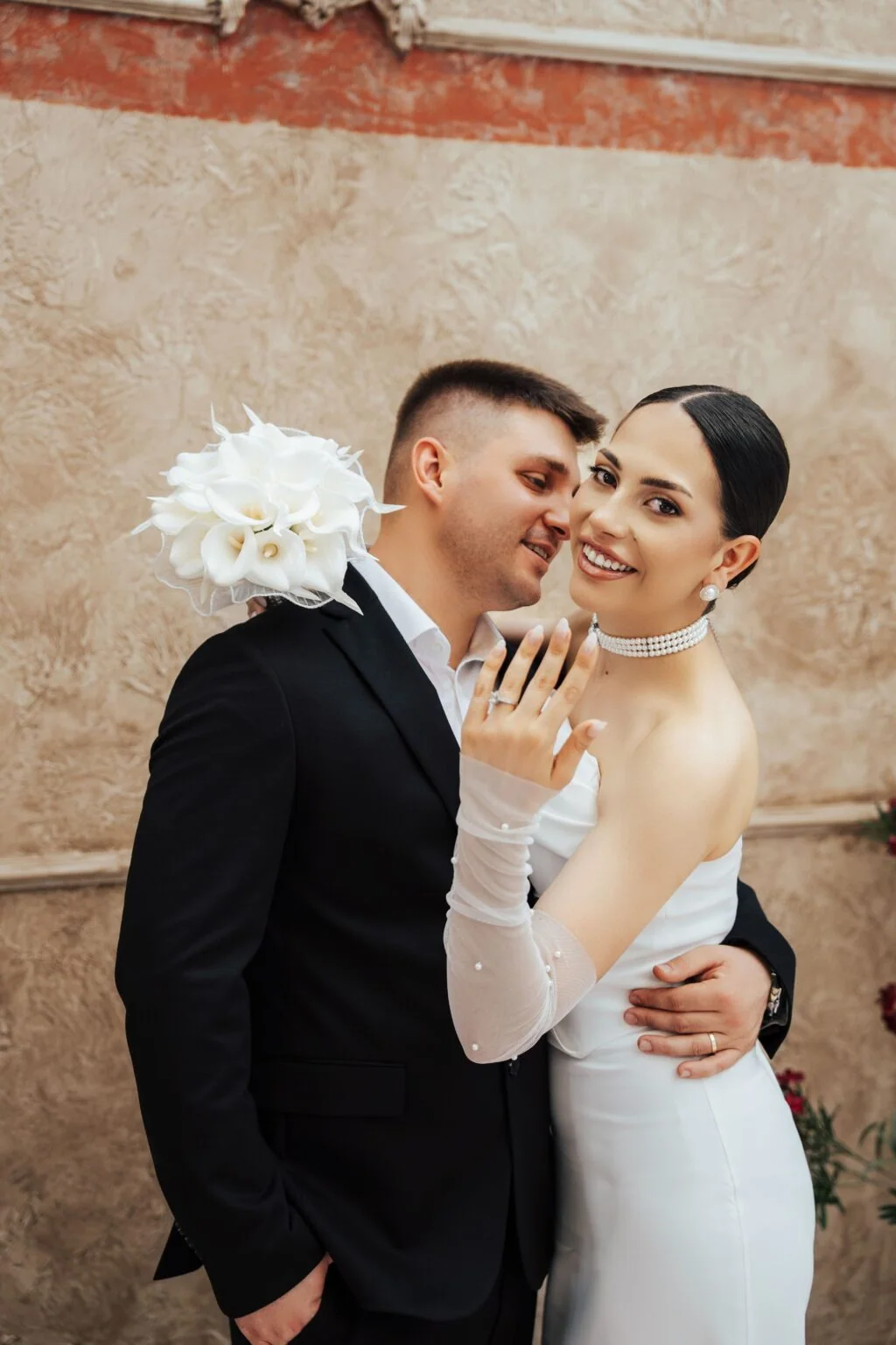 A couple embraces against a pale wall as the bride smiles toward the camera.