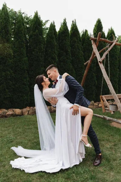 The groom carries the bride in front of a hedge and a wooden swing.