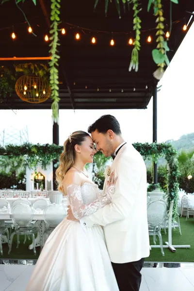 The newlyweds lean toward each other beneath a wooden structure with string lights.
