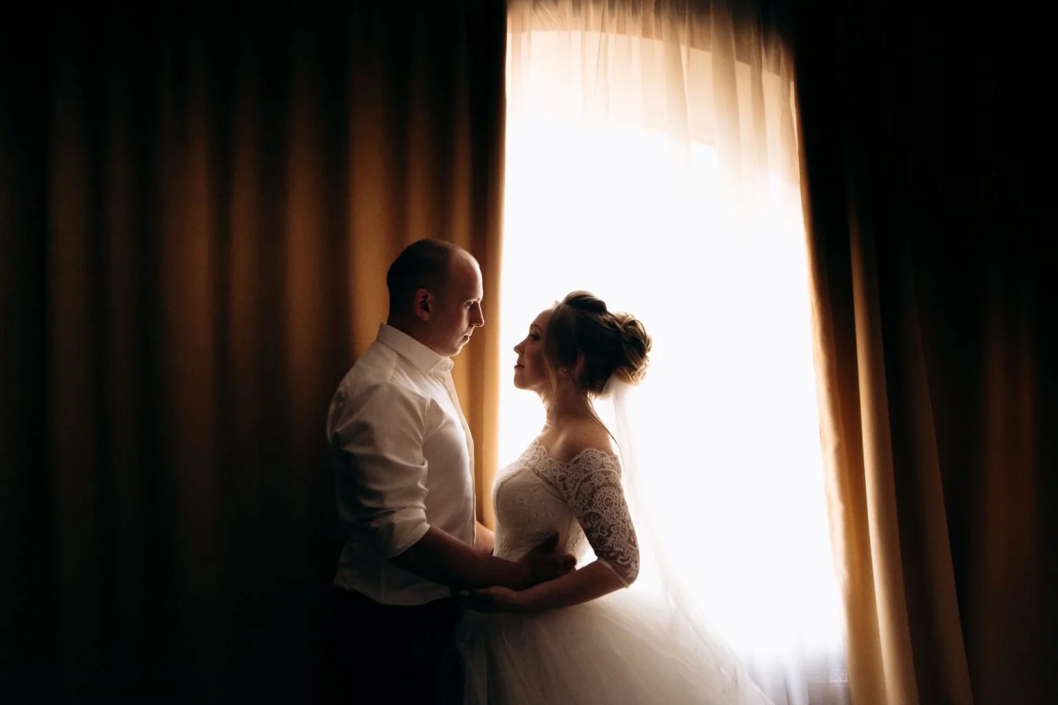 The newlyweds face each other in the light of a curtained window.